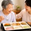 日本人の高齢女性が家族に見守られながら、やわらか食・ムース食・ペースト食などの介護向け宅配弁当を食べる様子