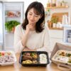 冷蔵庫・冷凍庫・食卓の宅配弁当を見比べる日本人女性が、冷蔵・冷凍・常温の宅配弁当の違いを比較している画像